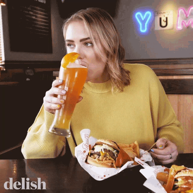 a woman in a yellow sweater is drinking a beer and eating a hamburger in front of a neon sign that says yum