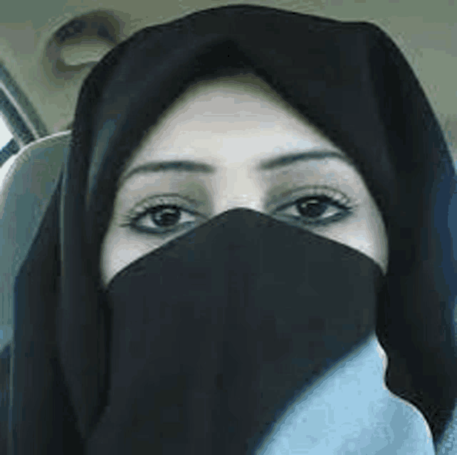 a woman wearing a black hijab is sitting in a car and covering her face with a black scarf .