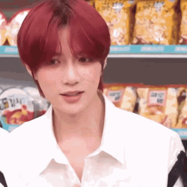 a young man with red hair is wearing a white shirt and standing in front of a shelf of chips .