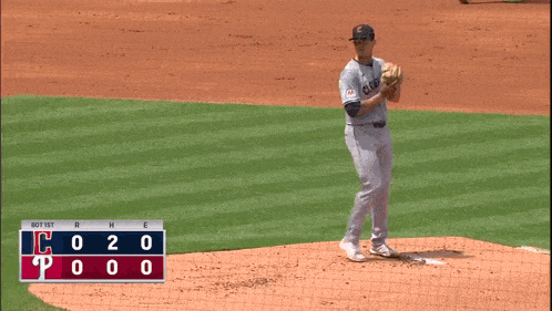 a baseball pitcher winds up to throw the ball with a scoreboard behind him that shows the score of the game