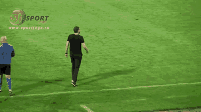 a man in a black shirt is walking on a soccer field with the website www.sportjugo.rs behind him