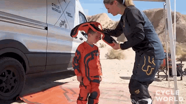 a woman is putting a helmet on a child 's head in front of a van that says cycle world