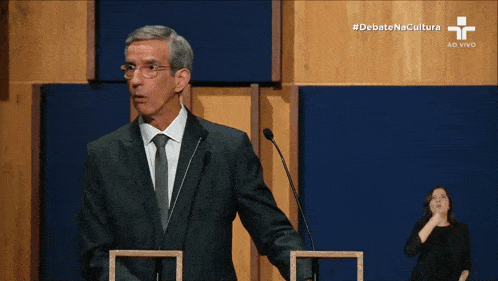 a man in a suit and tie stands at a podium in front of two microphones with debate na cultura written in the corner