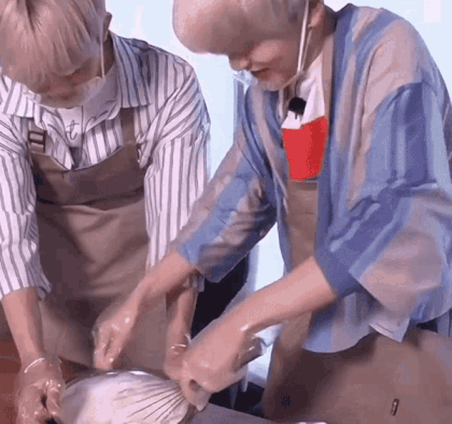 two men wearing aprons are preparing food on a table . one of the men is wearing a mask .