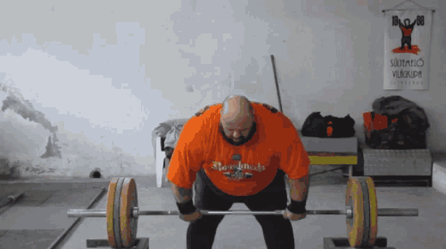 a man in an orange shirt is lifting a barbell in front of a sign that says " szombato vilokupa "