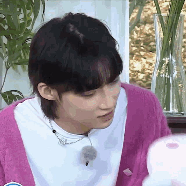 a young man wearing a pink cardigan and a white shirt is sitting in front of a vase of flowers .