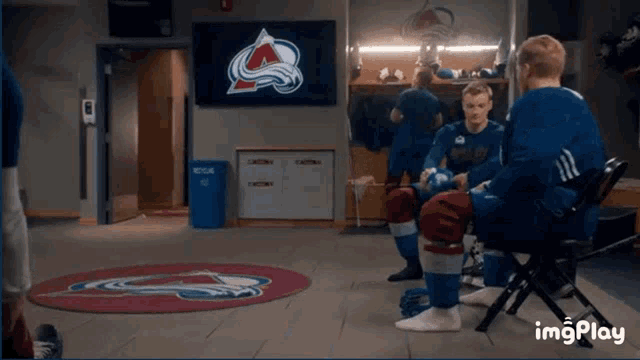 a group of hockey players are sitting in a locker room with a tv screen behind them that says avalanche on it