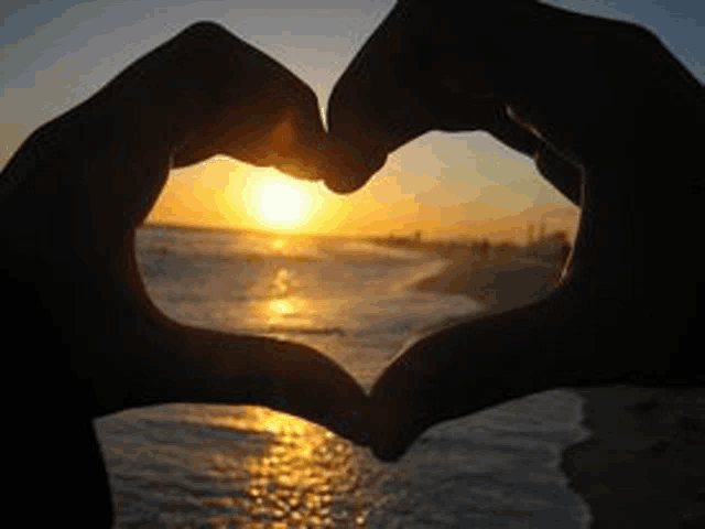 a person is making a heart shape with their hands on the beach at sunset .