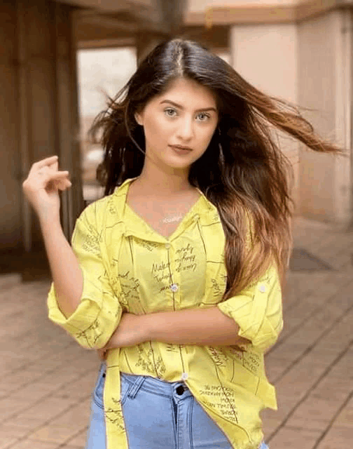 a young woman wearing a yellow shirt and blue jeans is standing with her arms crossed .