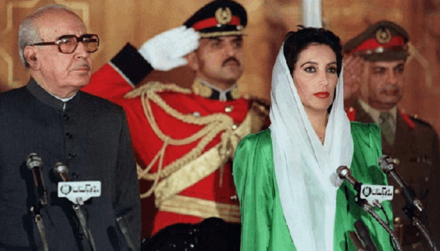 a man in a red uniform salutes while a woman in a green dress stands behind microphones