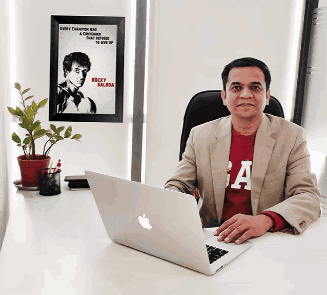 a man sitting at a desk with an apple laptop and a poster of rocky balboa