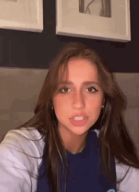 a young woman with long brown hair is sitting on a bed in front of a wall with frames .