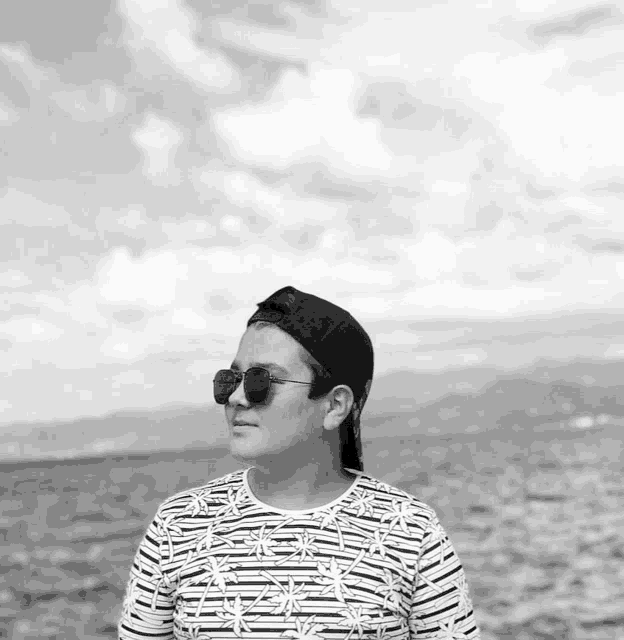 a boy wearing sunglasses and a baseball cap stands in front of the ocean