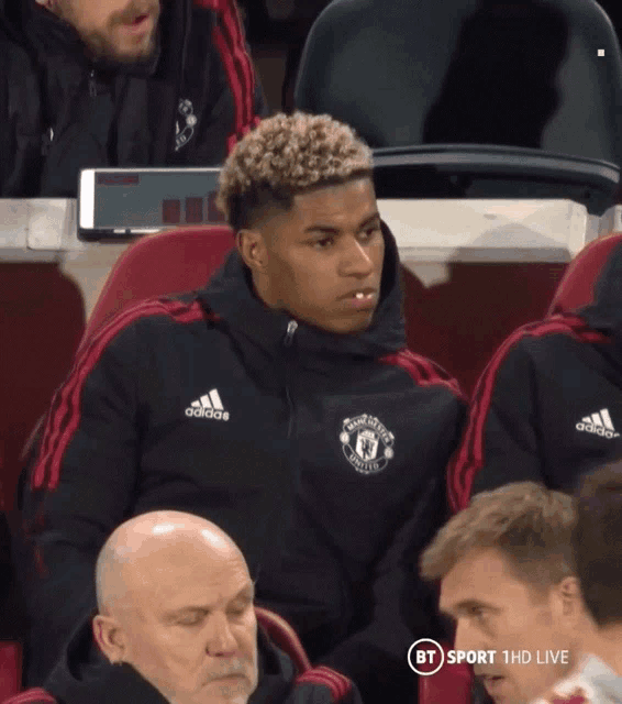 a man wearing an adidas jacket sits in a soccer stadium