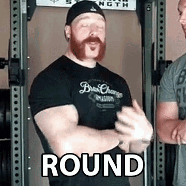 a man with a beard wearing a black shirt and a black hat is standing in front of a barbell rack .