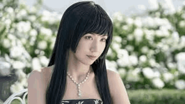 a woman with long black hair and a necklace is sitting on a bench in front of a bush of white flowers .