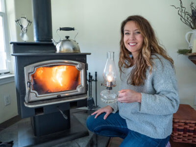 A woman crouched by a woodstove with a fire going, holding an oil lamp.