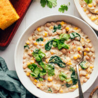 Spoon in a bowl of creamy vegan white bean chili next to cilantro and slices of cornbread