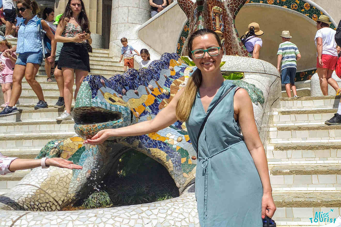 The writer of the post in a light blue dress poses next to a colorful mosaic lizard sculpture, with people walking down the stairs in the background.