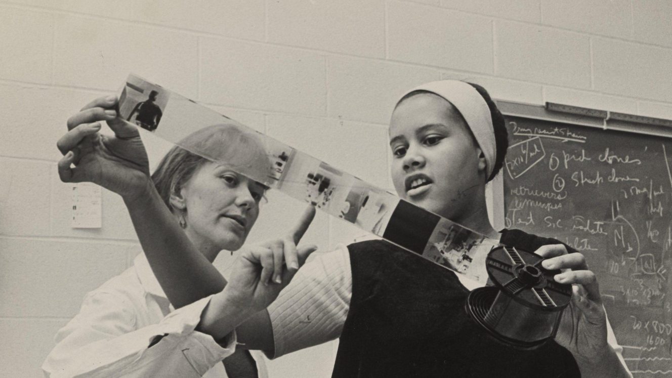 Black and white archival photo of Eva Irene Kataja pointing to a negative on a reel held by Toni Peters.