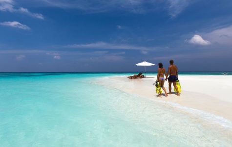 Two people walking on a white sandy beach with clear blue water, holding snorkeling fins.