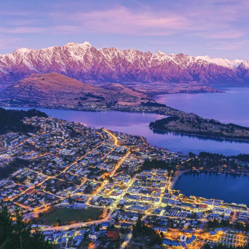 Twilight view of a lakeside town with illuminated streets against snow-capped mountains in New Zealand