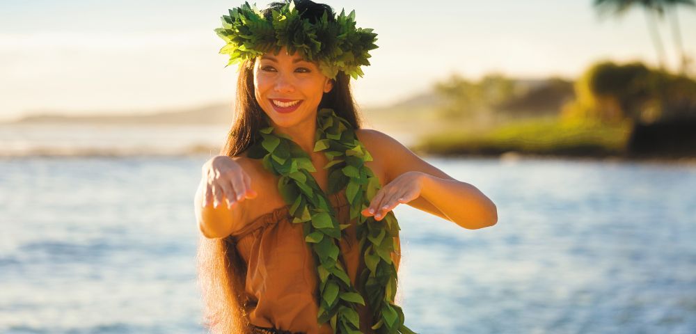 Person in traditional hula attire with a leaf skirt and headpiece by the water at sunset.