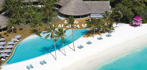 Aerial view of a tropical resort with a pool beside a white sandy beach and a pink lifeguard hut.