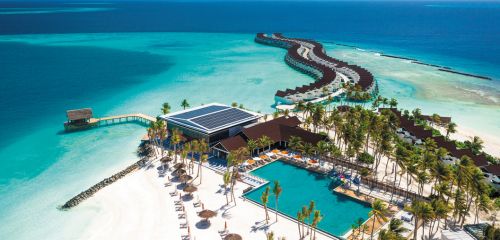 Aerial view of a tropical resort with overwater bungalows, a pool, and palm trees by a clear blue sea.