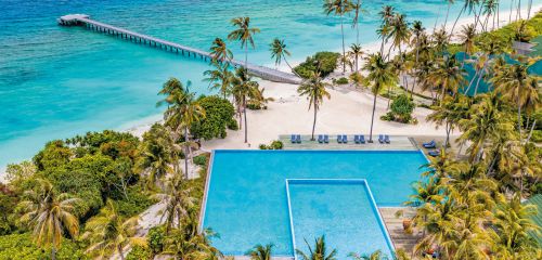 Aerial view of a tropical resort with a pool, palm trees, white sandy beach, and a pier extending into the ocean.