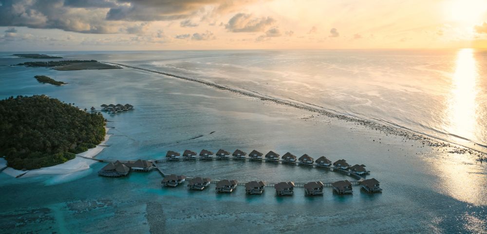 Overwater bungalows at sunset with calm sea and a sunlit sky.