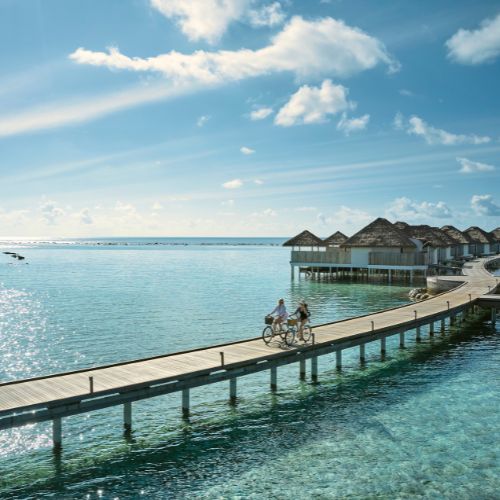 Two people cycling on a wooden pier with overwater bungalows and ocean view.