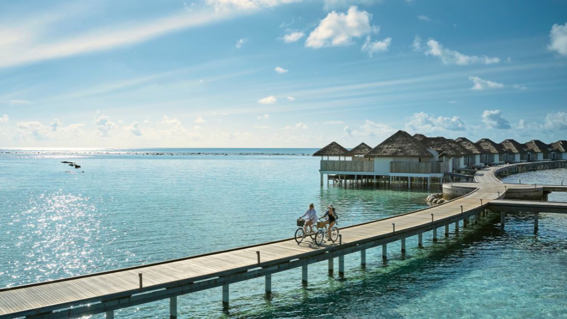 Two people cycling on a wooden pier with overwater bungalows and ocean view.