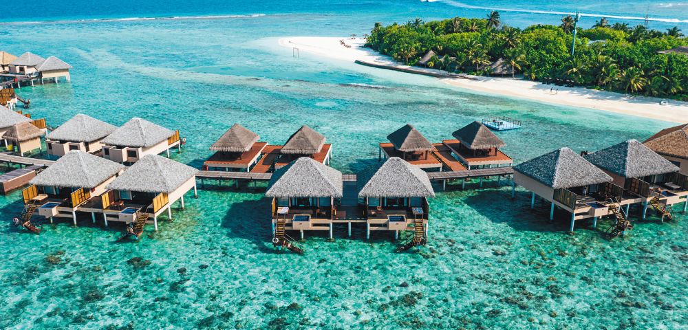 Overwater bungalows with thatched roofs in a clear turquoise sea near a tropical beach.