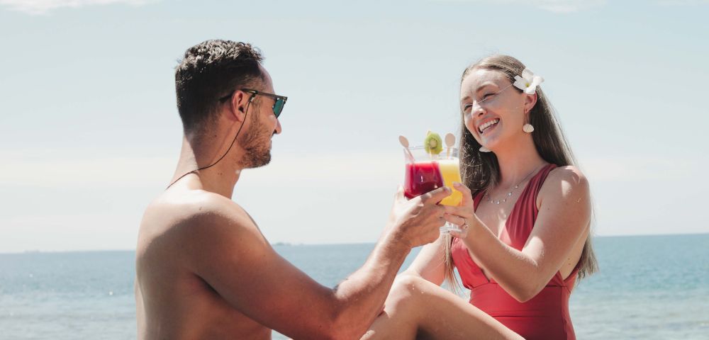 Two people toasting drinks on a sunny beach.