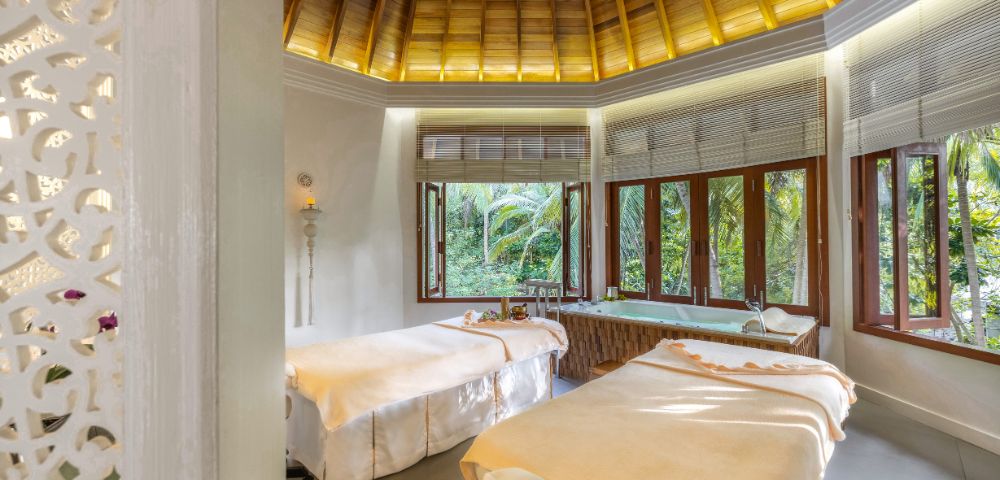 Luxury spa room with two massage tables, bamboo ceiling, and lush greenery visible through windows.