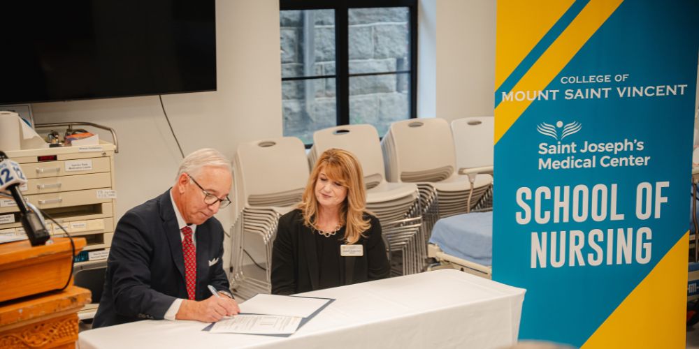 President and CEO of St. Joseph's Medical Center Mike Spicer signs an agreement alongside President of the University of Mount Saint Vincent Susan Burns, as they sit in a healthcare simulation laboratory with a banner next to them that says: University of Mount Saint Vincent Saint Joseph's Medical Center School of Nursing