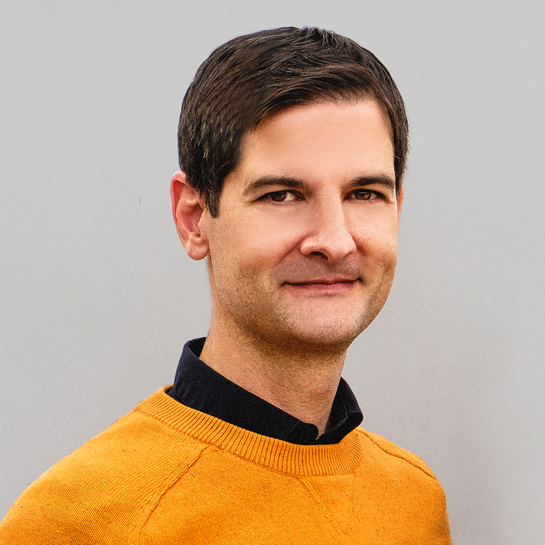 Photo of Joe Poindexter wearing a mustard colored sweater standing in front of a grey wall and smiling at the camera.