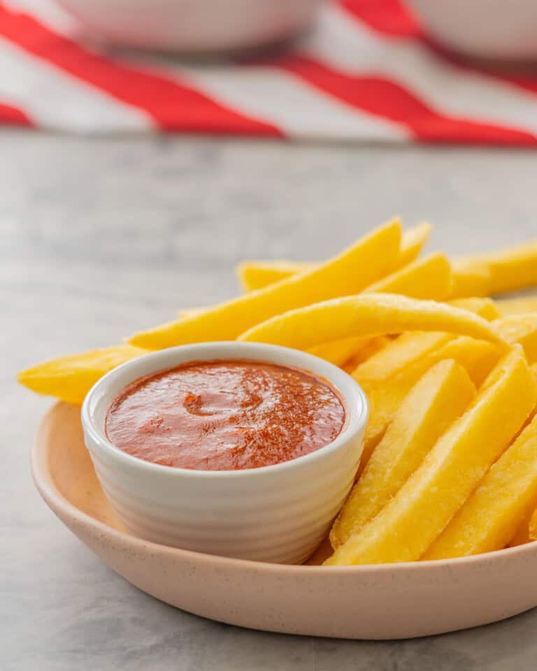 A bowl of fries with homemade ketchup in small ramekin inside bowl on benchtop with folded red and white stripped table cloth.