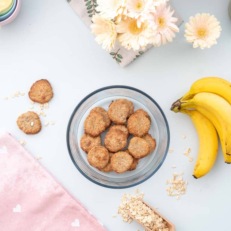 A round glass container full of oatmeal cookies on a bench next to a bunch of bananas.