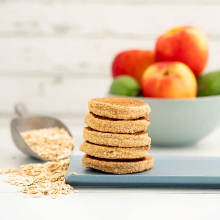 A stack of five pancakes on a blue platter, a bowl of apples and rolled oats in the background.