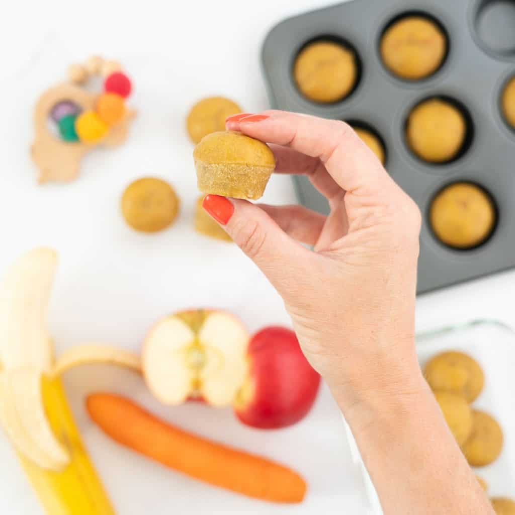 A women's hand holding a mini muffin above a tray of baked mini muffins.