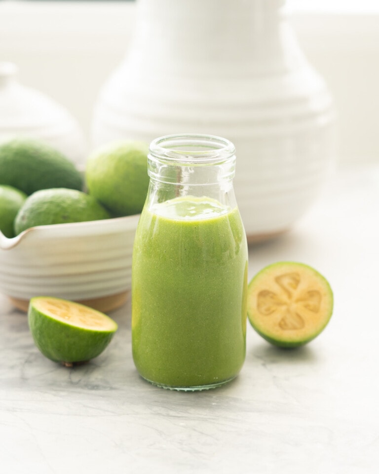 A small glass bottle of green smoothie with a green and white striped straw, a bowl of feijoas in the background.