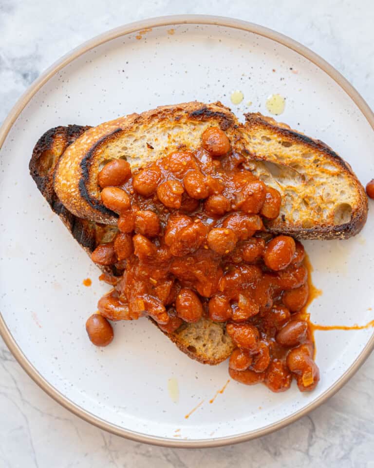 Two slices of rustic toast topped with baked beans on a white plate with brown rim.