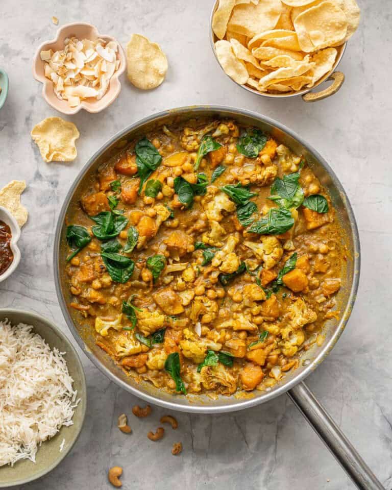 A skillet of chickpea and pumpkin curry next to a bowls of rice, poppadoms, relish, coconut flakes and cashews.