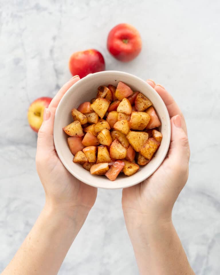 A bowl of cubed cooked cinnamon spiced apples being held above a marble bench top.