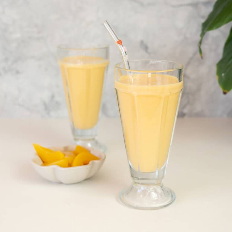 A tall sundae glass filled with a pale yellow smoothie with a pink straw, bowl of peaches in the background.