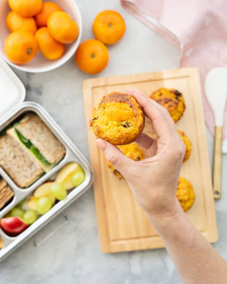 Hand holding a Mandarin muffin with rest on bench top with a lunchbox filled with mandarin muffin, sandwich, cherry tomatoes, muesli bar, apples and grapes.