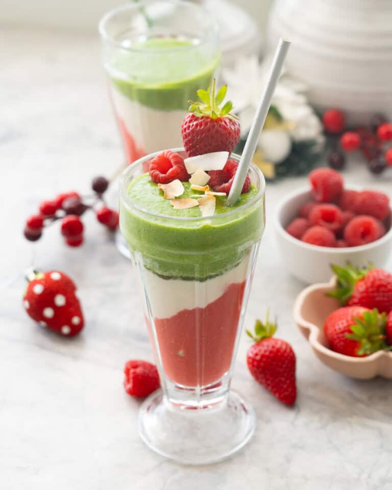 Tall cup with layered Christmas smoothies inside, red at the bottom, white in the middle and green smoothie at the top. Garnished with dried coconut flakes and strawberries and raspberries. Strawberries and raspberries on bench top and in small ramekins in background.