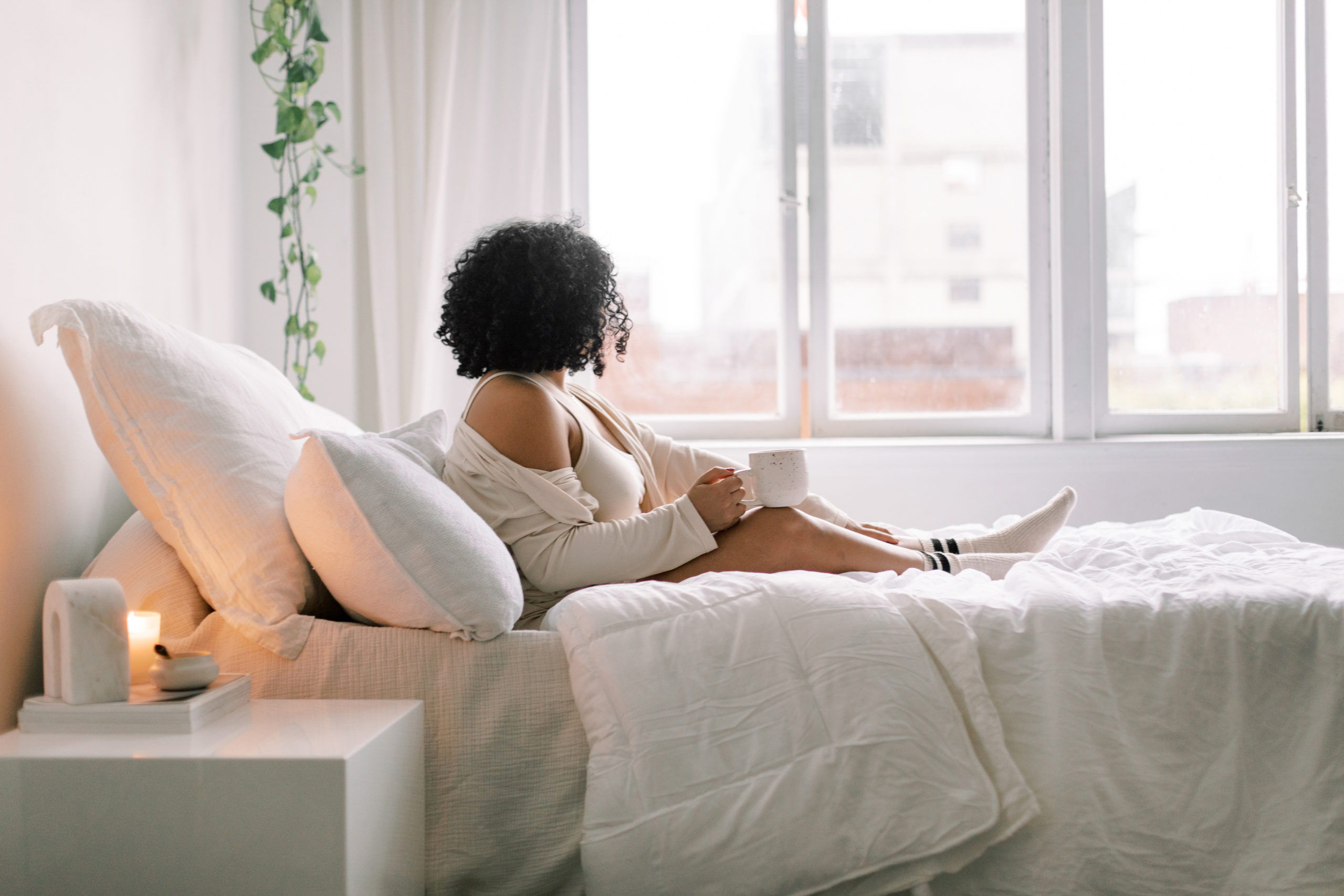 Woman sat on bed with a cup of coffee looking out the window thinking about how to create a positive environment for her self esteem.